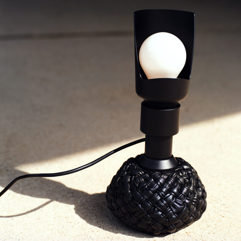 A modern black table lamp with a woven base and exposed light bulb is illuminated, casting shadows on a beige surface.