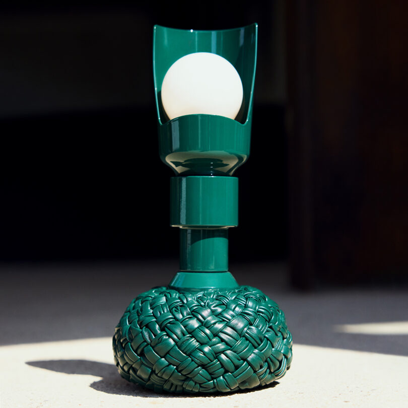 A green lamp with a woven base and a round bulb, illuminated in natural light.