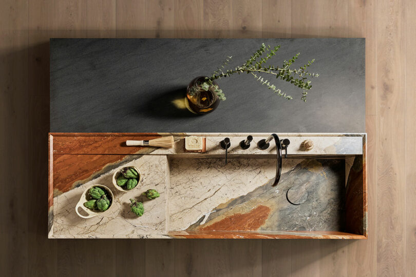 Top view of a modern kitchen counter with a marble finish, featuring a sink, faucet, cutting board, brush, two artichokes in a bowl, and a vase with greenery.