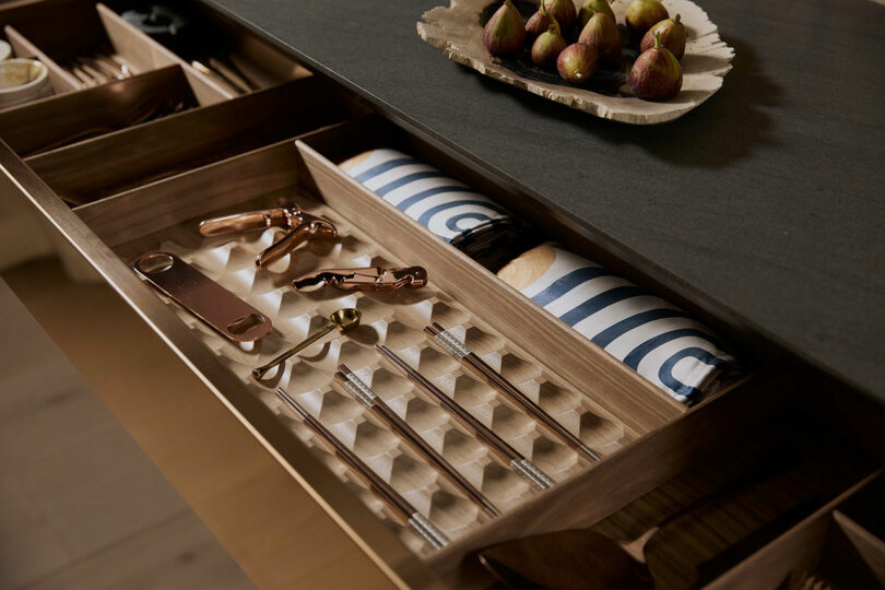 Open drawer with neatly organized utensils, including spoons and corkscrews, alongside a striped napkin and a set of figs on a tray above.