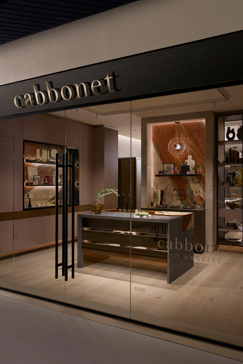 Showroom interior displaying a modern kitchen design with a central island, decorative shelving, and wood accents, viewed through a glass window labeled "cabbonet" in Los Angeles.