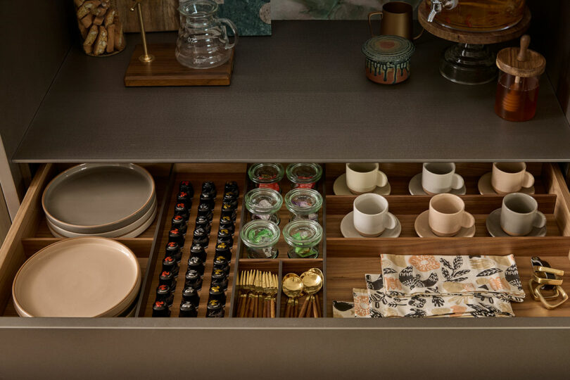 A drawer open to reveal organized tableware: stacked plates, cups, jars, and arranged cutlery next to napkins with a patterned design.