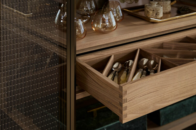 Open wooden drawer with compartments holding cutlery; above are glassware and spice jars on a tray.