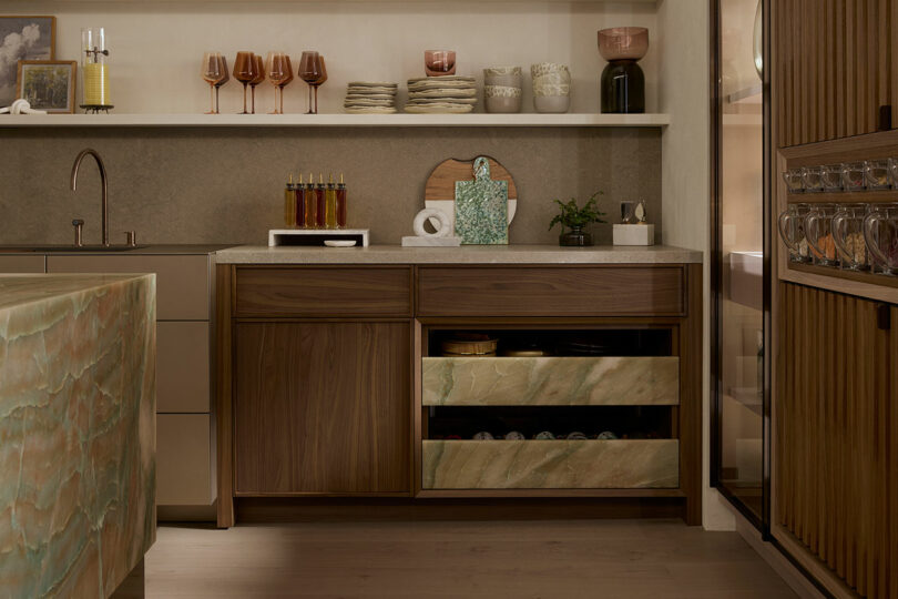 A modern kitchen with wood cabinets, open shelves, glassware, and a marble island. The lower cabinet drawers are partially open, showing stored items.