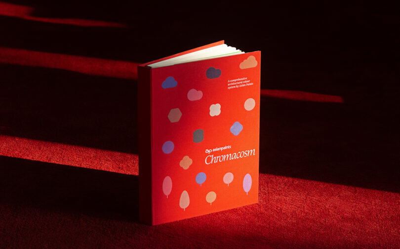 A red book titled "Chromacosm" with colorful abstract shapes on the cover, standing upright on a red surface with light and shadow patterns.