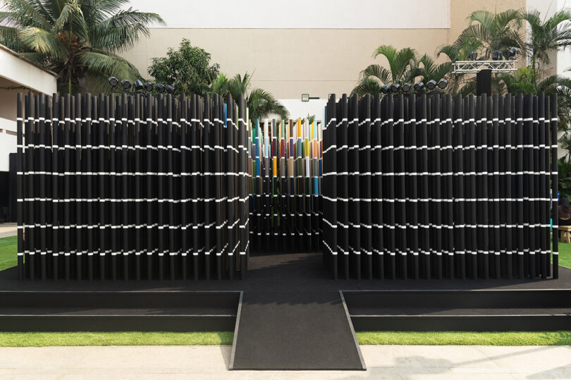 An outdoor installation featuring two rows of black vertical panels with white stripes, leading to a colorful section in the center, surrounded by greenery.