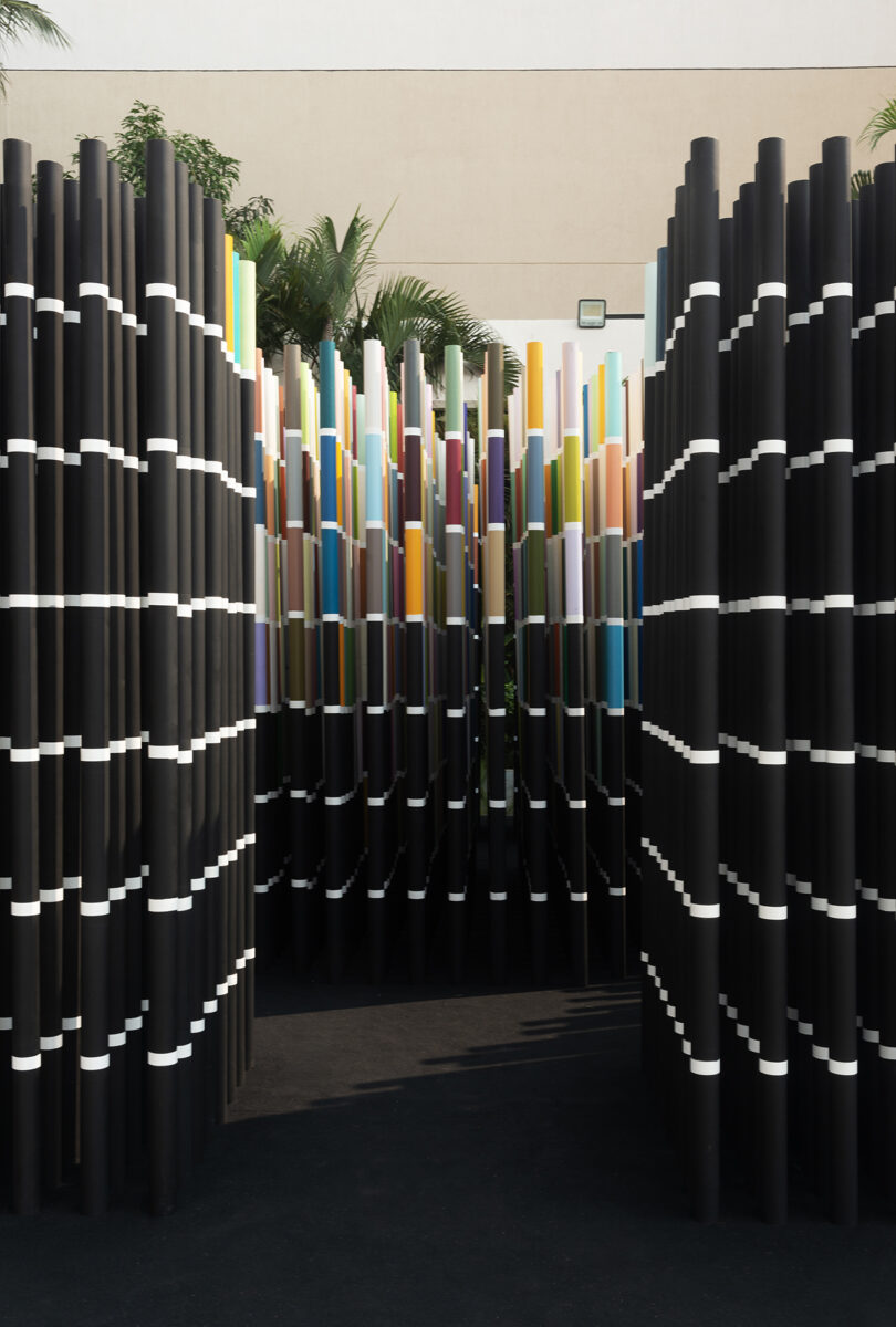 Installation of tall, black vertical rods with colorful tops, arranged in two rows, creating a narrow pathway. Background includes a building wall and green foliage.