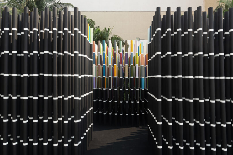 Installation of vertical black tubes with white bands, forming a passageway, leading to a colorful section at the center.