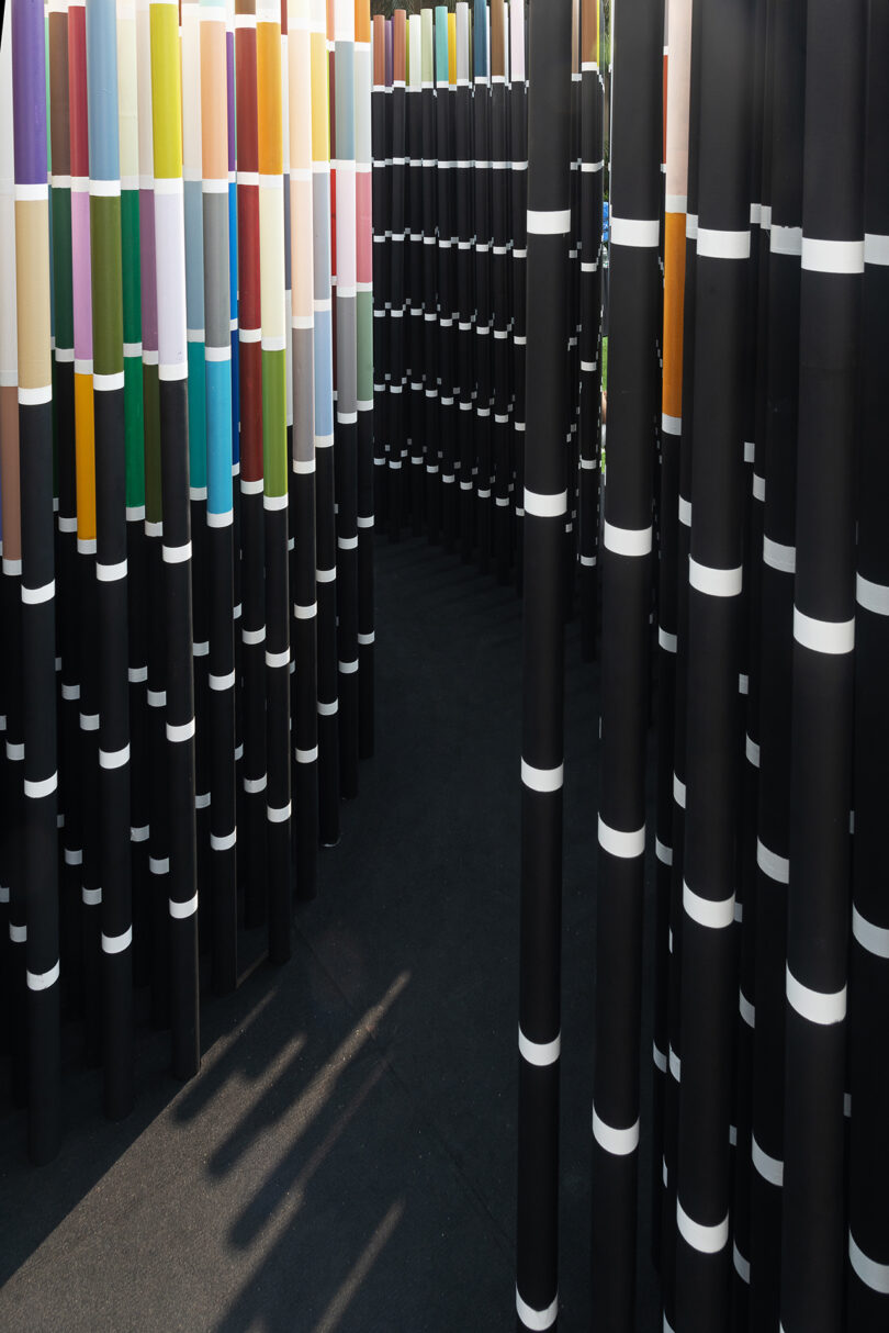 An installation of tall, black cylindrical structures with horizontal white bands, interspersed with colorful pillars, creating a maze-like pathway.