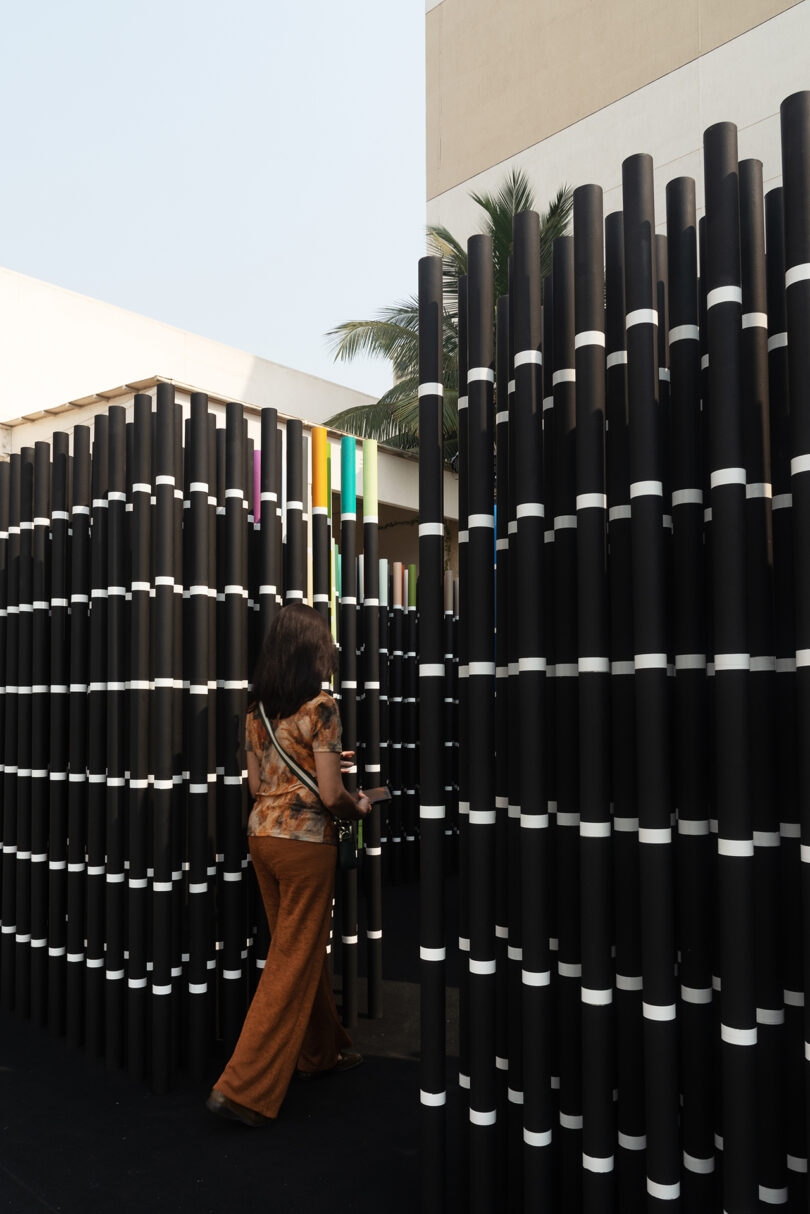 A person walks through an outdoor installation of vertical black poles with white bands, positioned in rows.