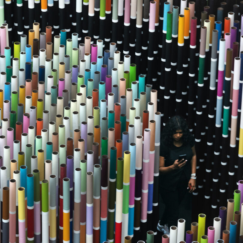 A person stands amidst a dense arrangement of colorful vertical tubes, looking at a smartphone.