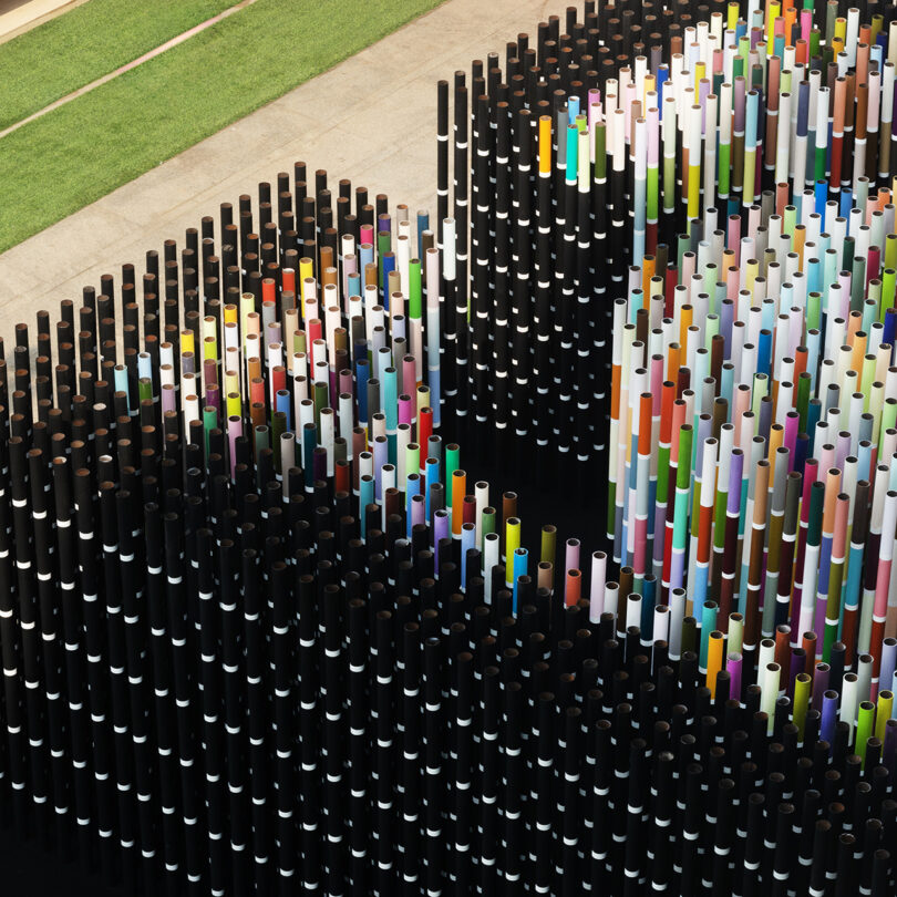 Aerial view of a colorful art installation featuring numerous vertical tubes arranged densely in a pattern, situated near a patch of grass.