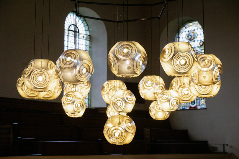 A cluster of spherical pendant lights with circular patterns, reminiscent of a CDW 2025 Product Awards nominee, hangs from the ceiling in a room graced by two stained glass windows.