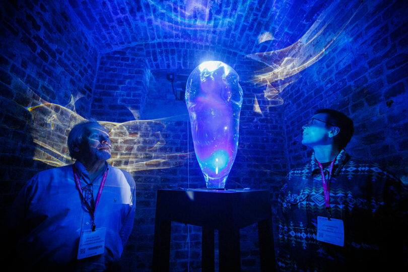 Two people admire a glowing blue sculpture on a pedestal in the dimly lit, brick-walled room, reminiscent of an exhibit worthy of the CDW 2025 Product Awards.