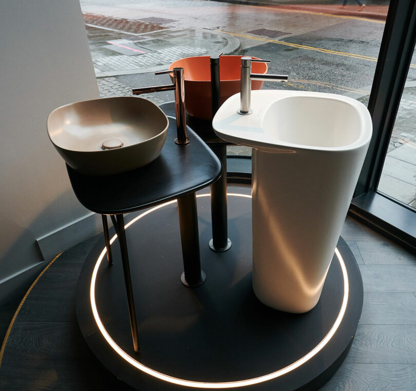 Three modern sinks on display, featuring two vessel sinks on a black table and a freestanding sink beside them, all showcased near a large window overlooking the street. These designs are contenders for the prestigious CDW 2025 Product Awards.