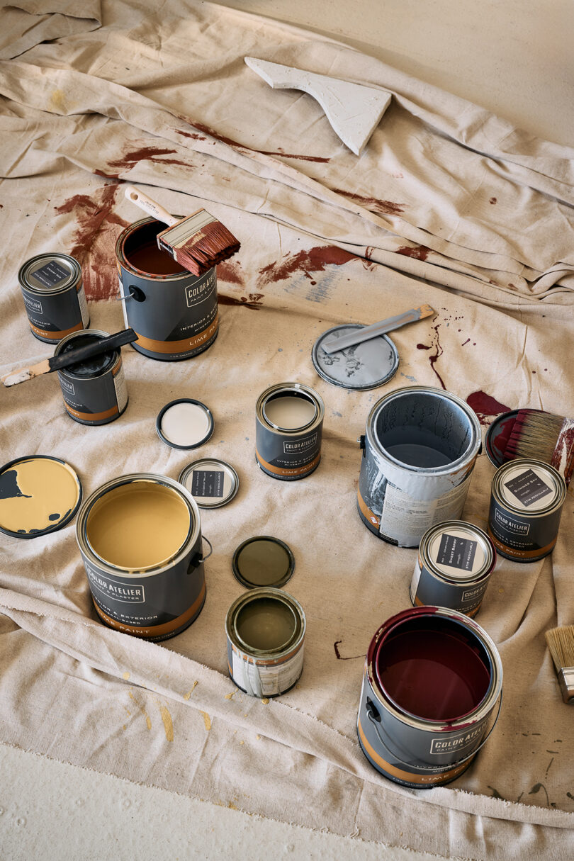 A collection of open paint cans with various colors on a drop cloth, accompanied by paintbrushes and a roller. Some paint has spilled onto the cloth.