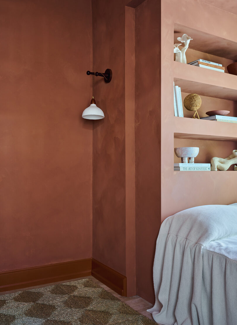 A cozy room with earth-toned walls, a wall-mounted light, a textured carpet, and built-in shelves holding books and decorative items. A light-colored blanket drapes over a surface.