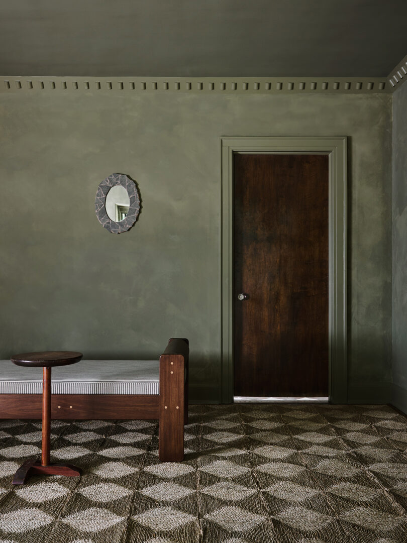 Room with green walls featuring a wooden door, mirror, bed with striped bedding, circular wooden side table, and patterned carpet.