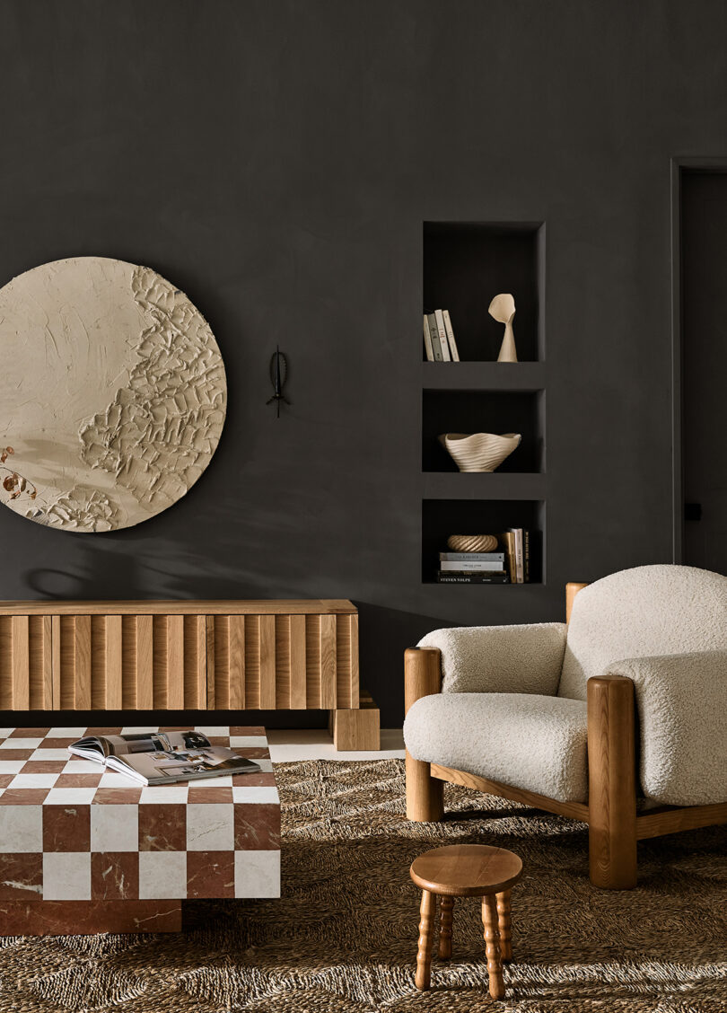 A stylish living room with a textured beige armchair, a checkered coffee table, wooden furniture, a round textured wall art piece, and built-in shelves with decorative items on a dark wall.