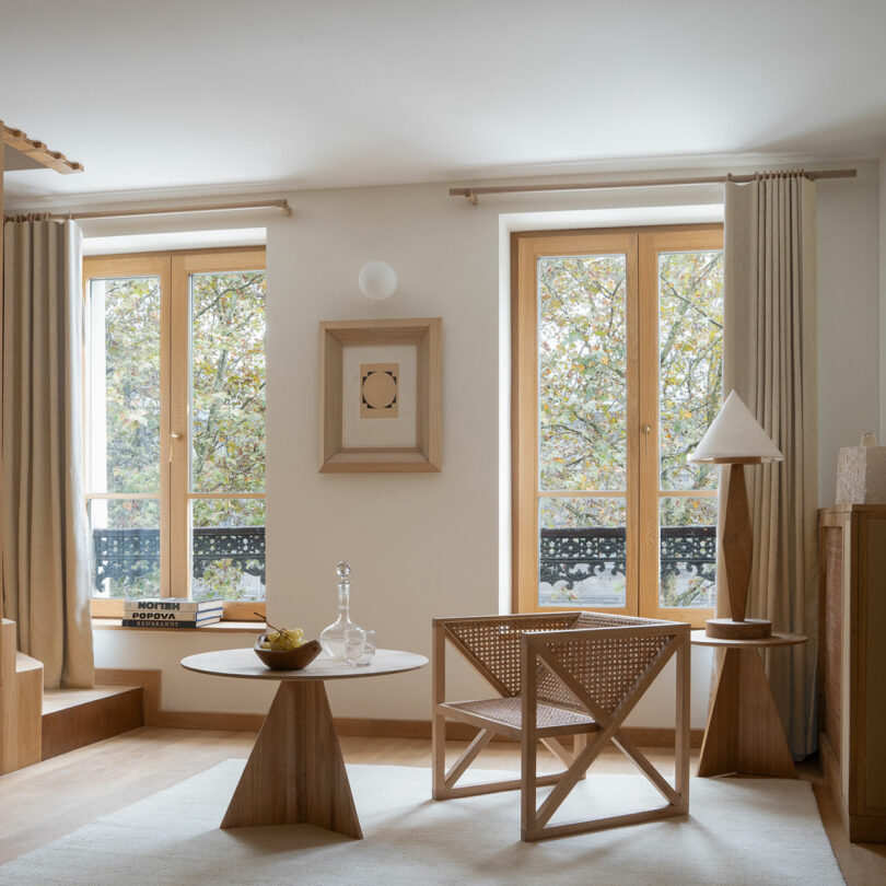 A minimalist living room with wooden furniture, a round table, a geometric chair, a tall lamp, and framed art exudes effortless elegance. Two large windows with beige curtains reveal an outdoor view of trees, creating a serene policronica scene.