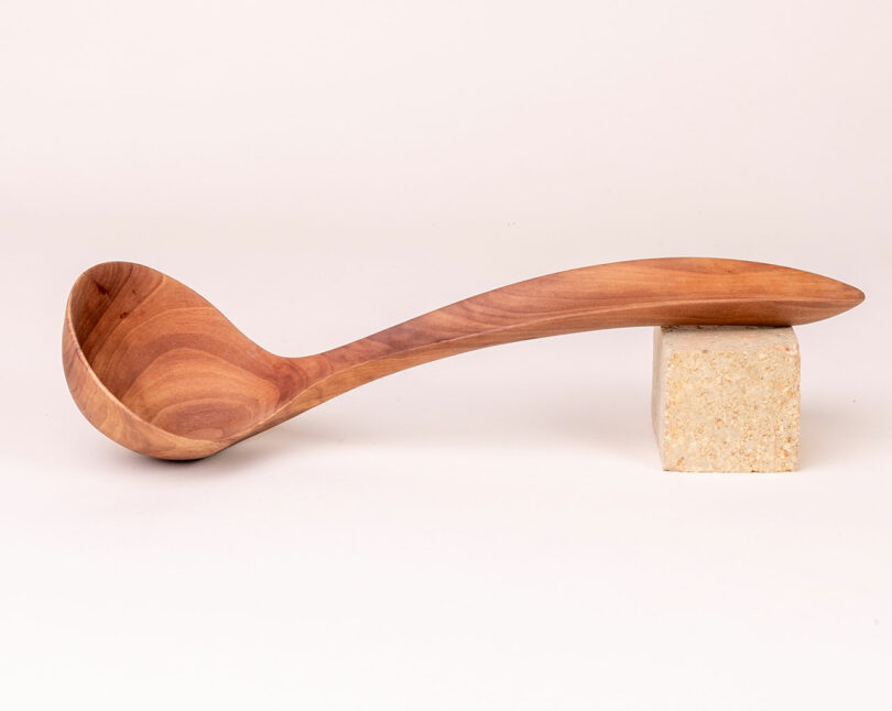 A smoothly carved wooden ladle, reminiscent of Alex Matisse's artistry, with a long, curved handle rests on a light-colored rectangular stone block.