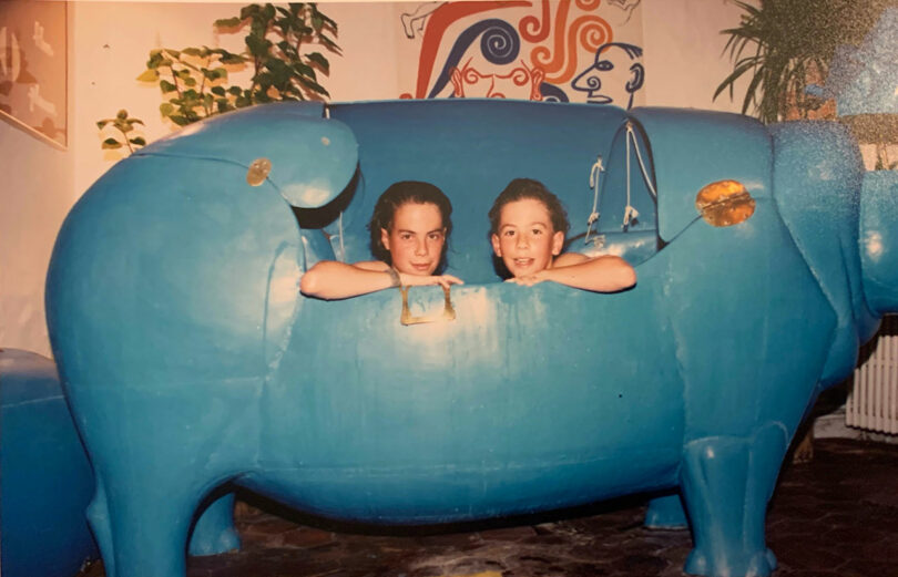 Two children sit inside a large blue hippo-shaped bathtub, reminiscent of an Alex Matisse sculpture. The room is adorned with vibrant plants and a colorful wall mural.