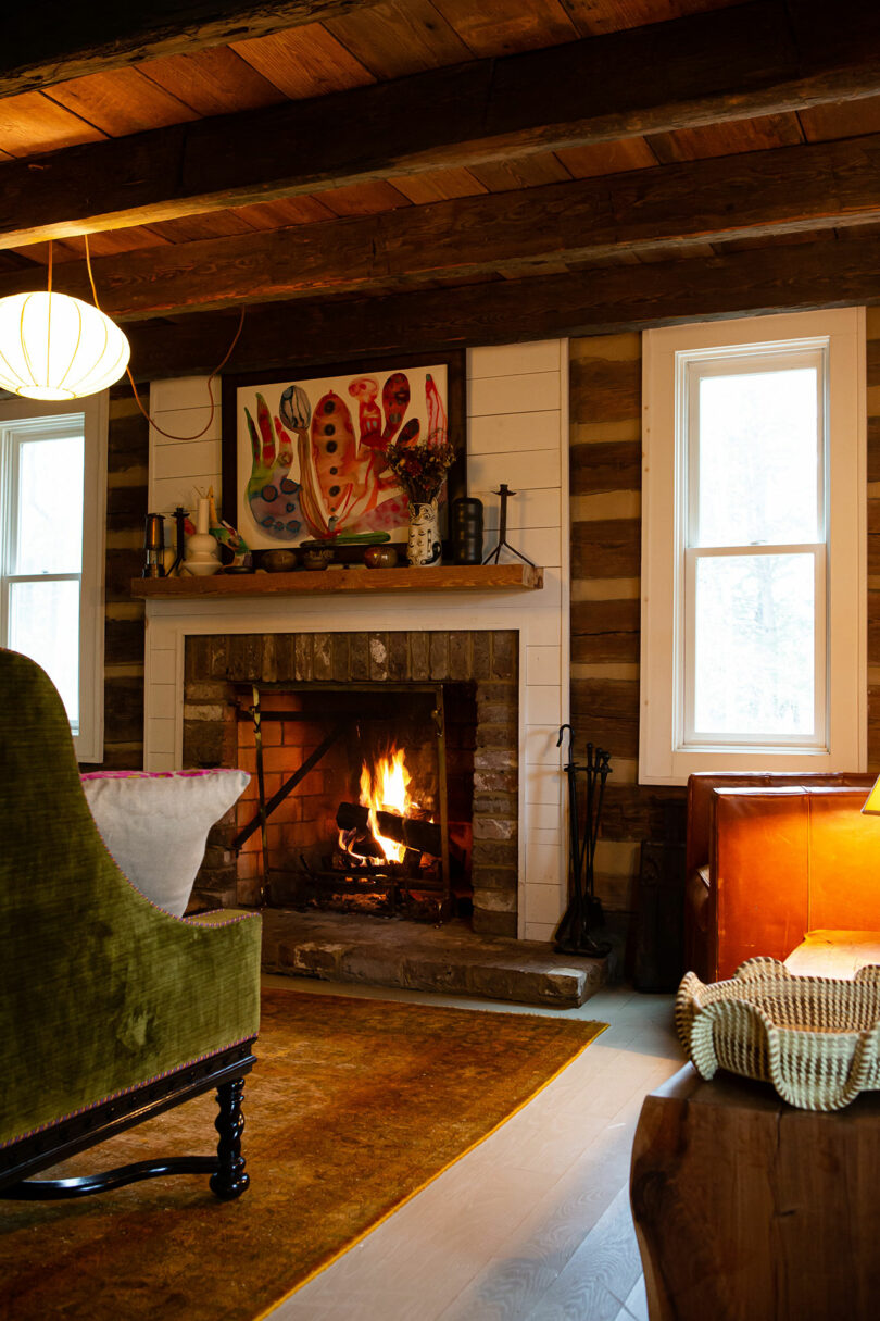 A cozy living room with a lit fireplace, wooden walls, and an Alex Matisse piece above the mantel. A green chair completes the scene, flanked by two windows that enhance the colorful painting. Soft lighting adds warmth to this inviting space.