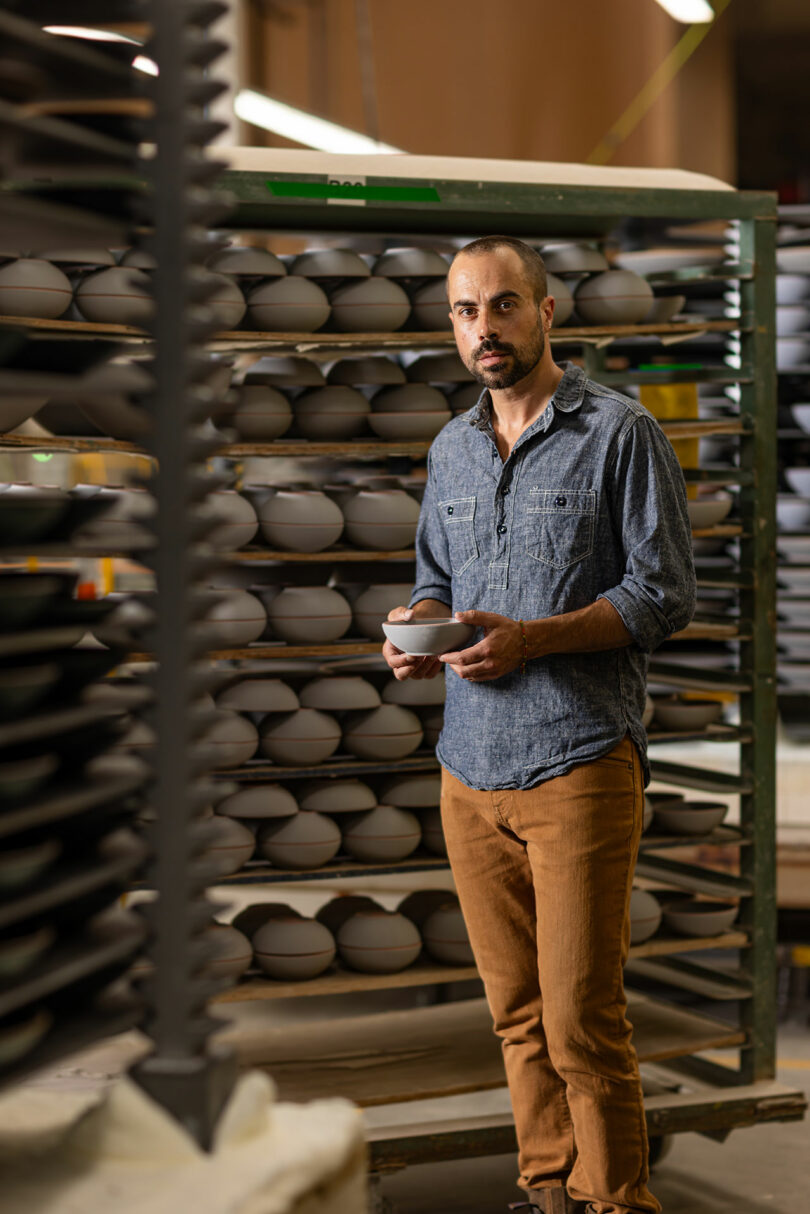 A man in casual attire stands in front of shelves filled with ceramic bowls, holding one crafted by Alex Matisse in his hands.