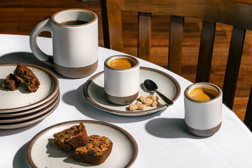 Coffee cups, plates with sliced cake, and a spoon rest elegantly on a white tablecloth, evoking the simple charm of an Alex Matisse design. In the background, a wooden chair complements the artisanal scene.