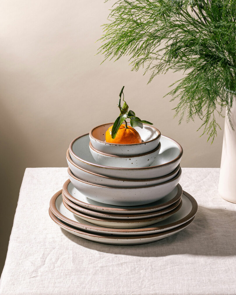 A stack of ceramic plates and bowls, reminiscent of Alex Matisse's craftsmanship, sits elegantly on the table with a small fruit perched on top. Nearby, a vase with lush green foliage complements the scene.