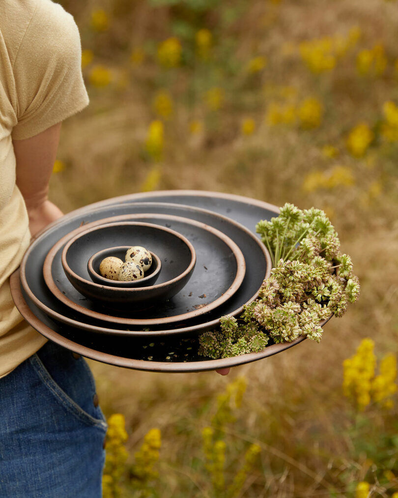 Person holding a stack of nested black plates with quail eggs in the smallest bowl and white flowers on the side, standing in a grassy field. The arrangement recalls the rustic elegance often seen in Alex Matisse's pottery designs.