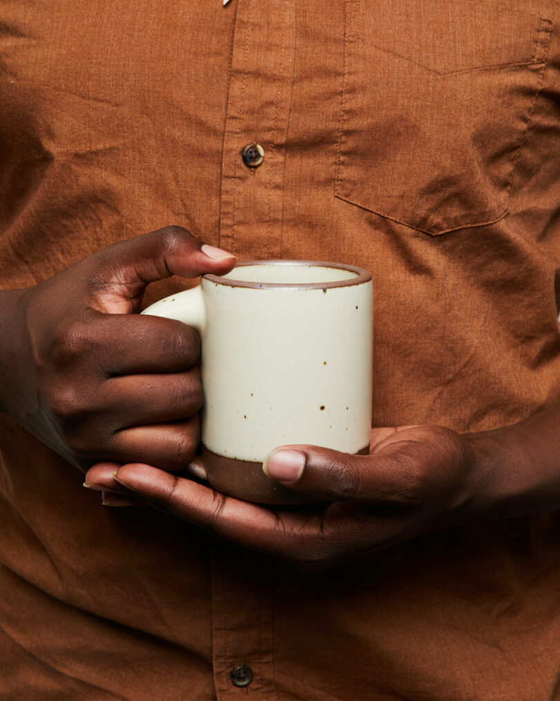 Holding a white ceramic mug with both hands, the person in a brown shirt evokes the artistry of an Alex Matisse creation.