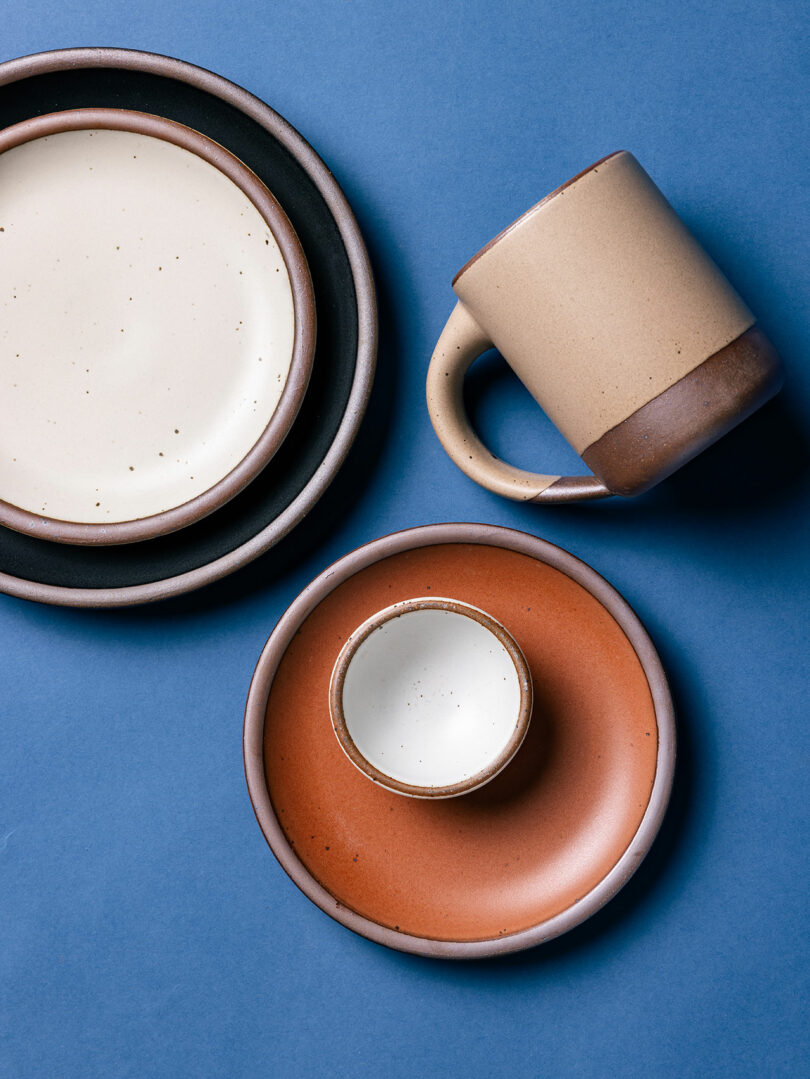 This assorted ceramic dinnerware set, reminiscent of Alex Matisse's artistry, features plates, a mug, and a cup arranged elegantly on a blue background.