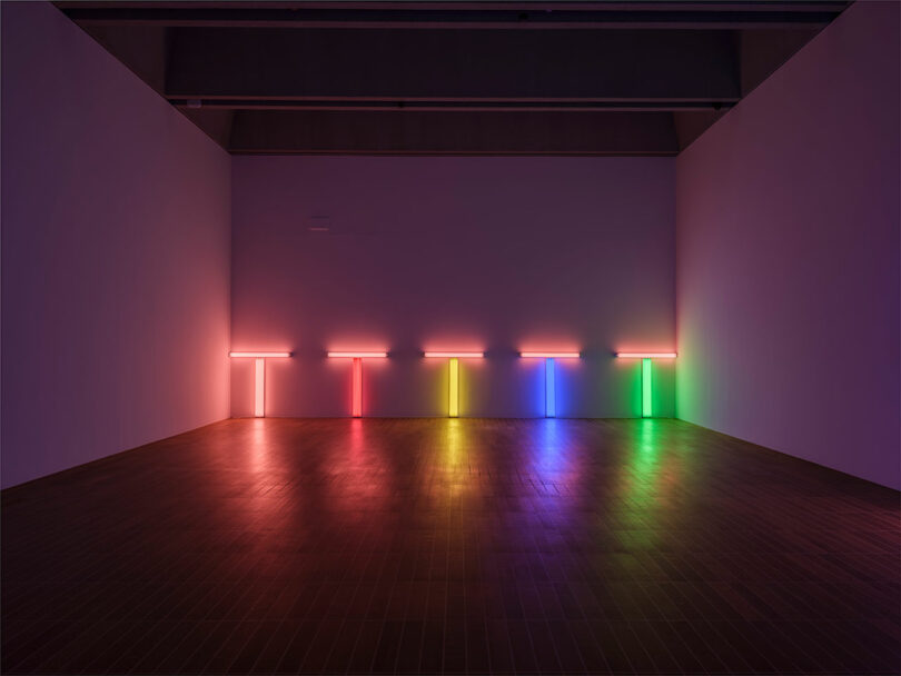 The dimly lit room, curated by Draga & Aurel, showcases horizontal neon lights in red, pink, orange, yellow, blue, and green. These vibrant hues are evenly spaced along three adjacent walls.