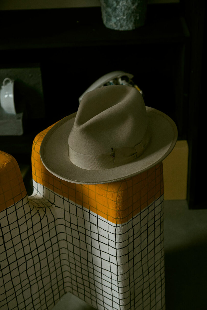 Un chapeau Fedora Taner avec un groupe de la collection Draga se trouve sur le dossier d'une chaise avec un motif à vérification caractéristique Aurel noire et orange, qui est placé devant un fond sombre.