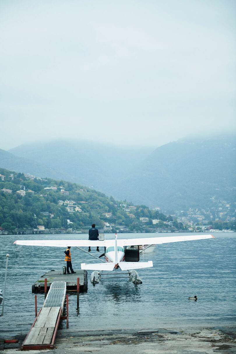 A seaplane docked by a wooden pier on a lake, with Draga and Aurel standing near the aircraft. The hilly landscape looms in the background under a cloudy sky.