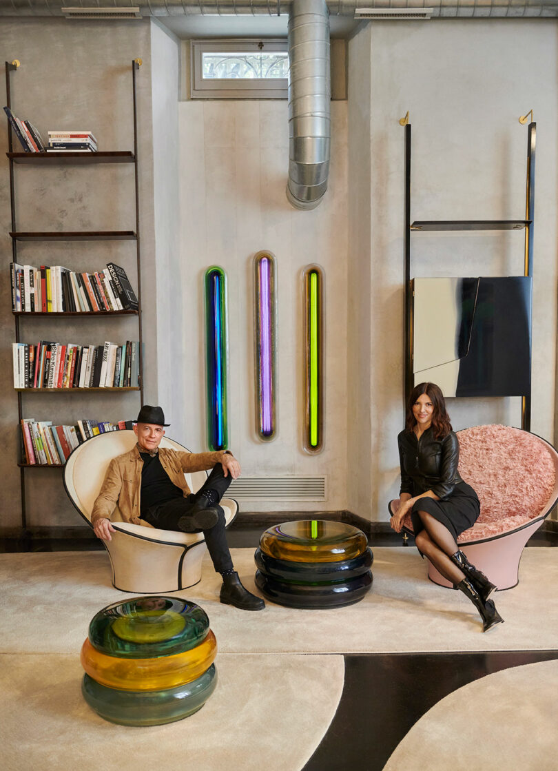 Two people sit on modern chairs in a stylish room adorned with colorful cylindrical lights, bookshelves, and contemporary tables. The sophisticated design evokes the signature elegance of Draga & Aurel's renowned aesthetic.