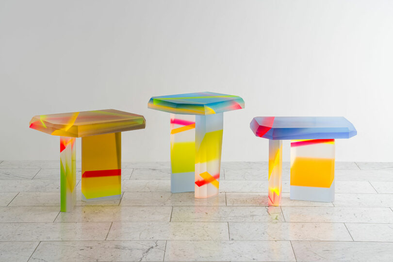 Three colorful, geometric acrylic tables with abstract patterns, designed by Draga & Aurel, are placed on a light tiled floor against a plain white wall.