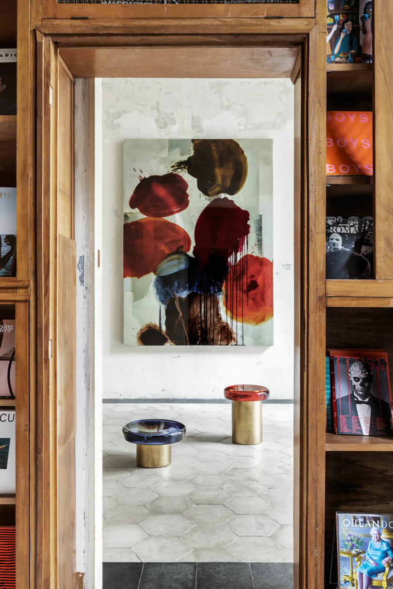 View through a doorway showing abstract art on a wall and two round stools. Shelves on either side hold various magazines, with a hint of Draga & Aurel's signature style subtly enhancing the ambiance.