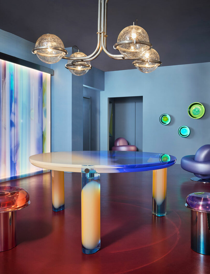 The modern room features a glossy, round table with blue gradient edges and cylindrical legs designed by Draga & Aurel. Futuristic chairs complement abstract wall art, while a striped backlight panel and a spherical chandelier add an extra touch of elegance.