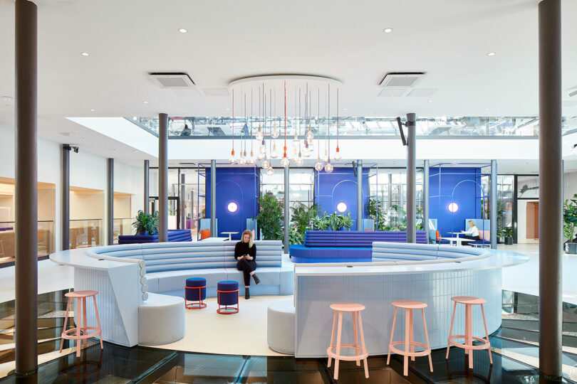 Modern, spacious interior with circular seating, blue accents, hanging lights, and a skylight. A person sits on the curved seats surrounded by greenery and bar stools.