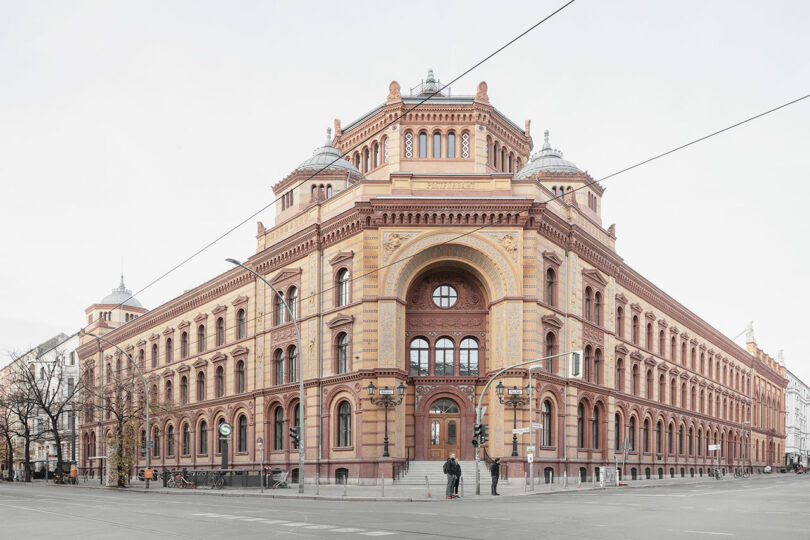 Historic red brick building on a corner with arched windows and decorative details, featuring four corner towers and an ornate entrance. Two people stand outside and tram lines cross the street.