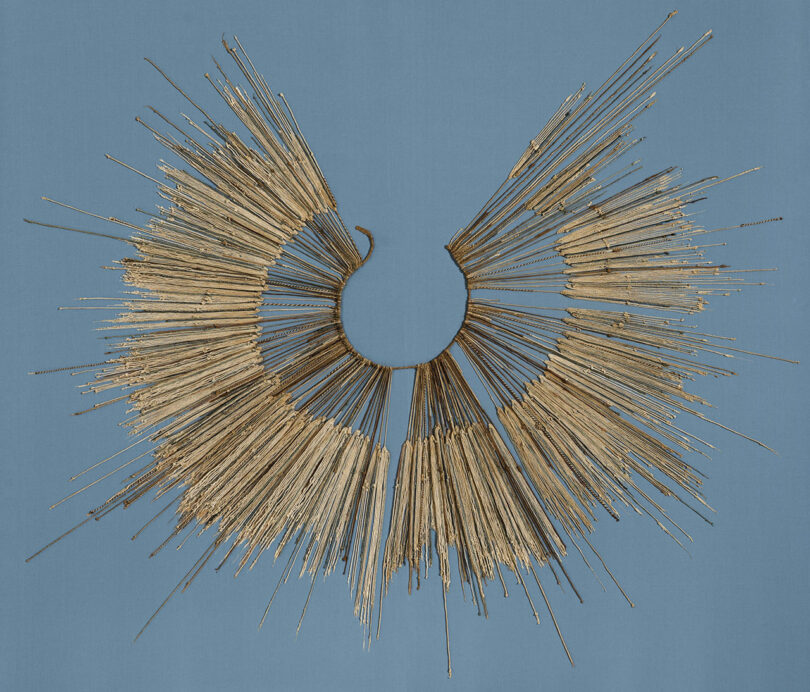 An ornate golden halo-like necklace, reminiscent of Rikke Frost's elegant designs, features numerous elongated spikes arranged in a circular pattern, beautifully displayed on a rich blue background.