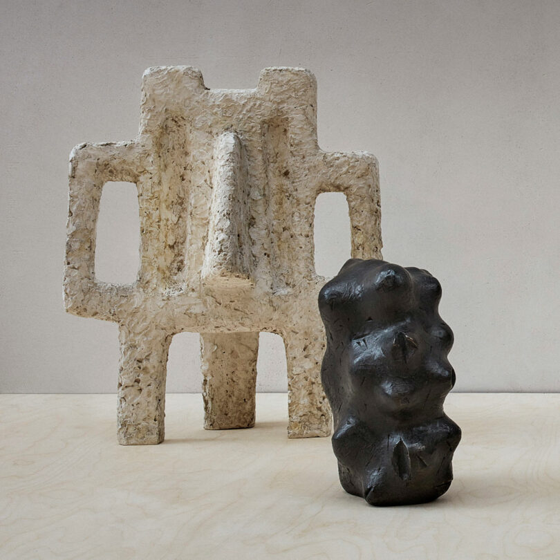 Two abstract sculptures: a tall, white, textured structure with geometric openings, reminiscent of Rikke Frost's unique style, and a shorter, smooth, dark form with irregular contours are placed on a light surface against a plain backdrop.