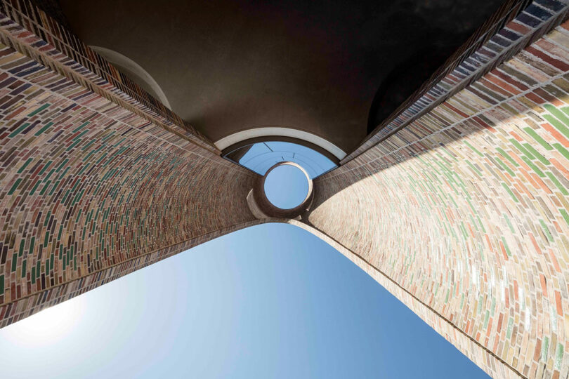 View looking up between two tall brick structures with a circular opening at the top, revealing the sky, reminiscent of Rikke Frost's architectural elegance.