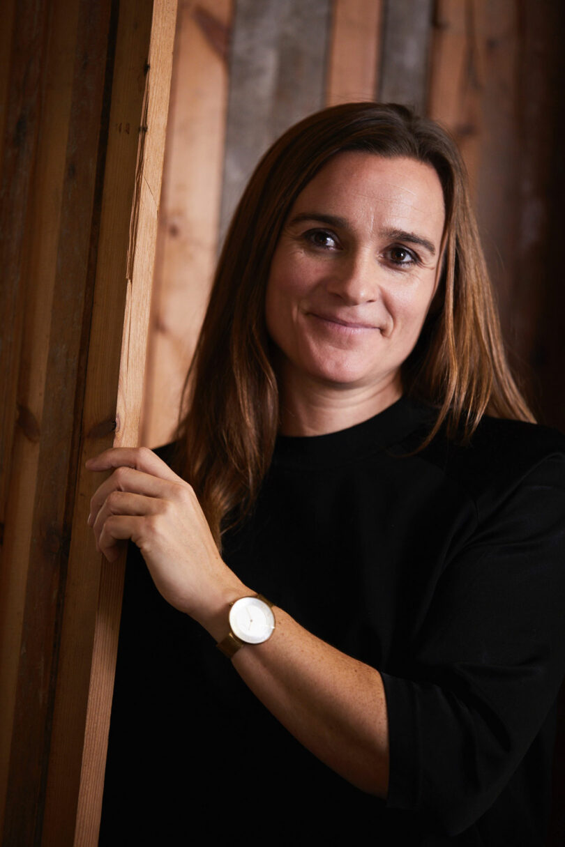 A person with long hair, wearing a black shirt and watch, stands indoors, gently holding a wooden beam while smiling slightly—a scene reminiscent of Rikke Frost's elegant style.