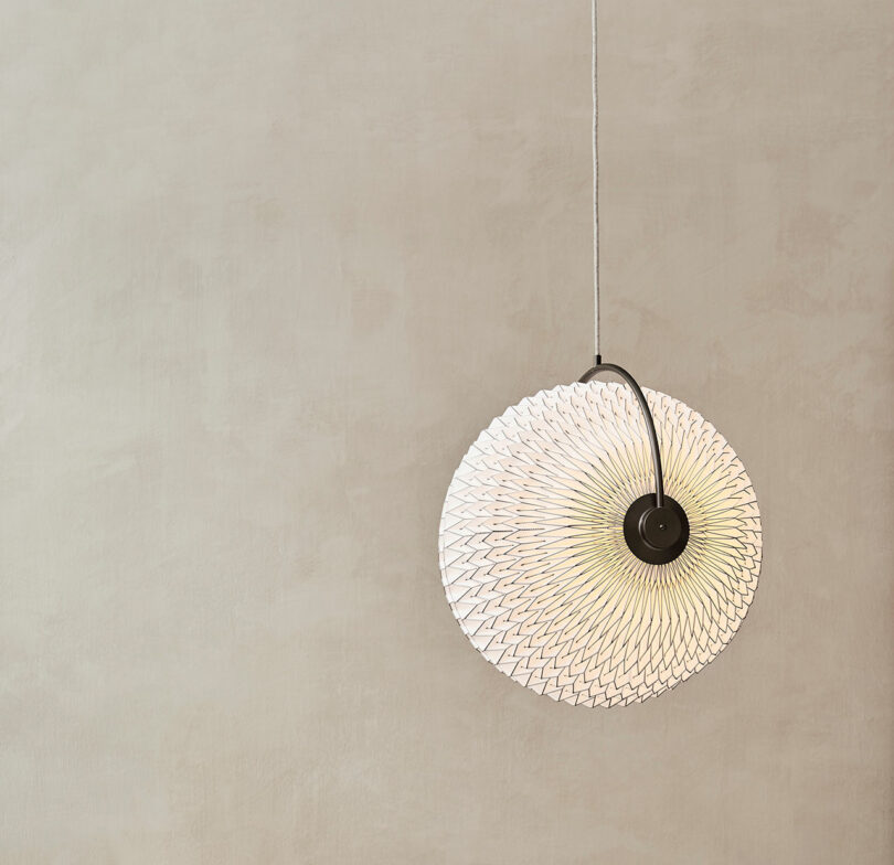 A round, textured pendant light, reminiscent of Rikke Frost's signature Scandinavian design, hangs against a plain beige wall.