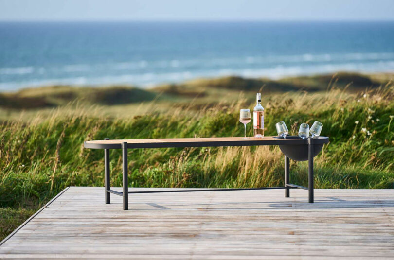 Outdoor table with two glasses, a bottle, and a small bowl on a wooden deck near grassy dunes, embodying the minimalist elegance reminiscent of Rikke Frost's designs, all overlooking the serene ocean.