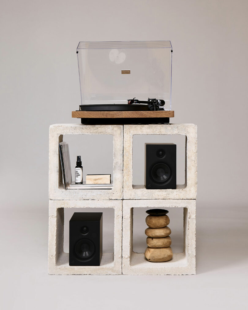 A Gadhouse turntable rests on cinder blocks surrounded by sleek speakers, vinyl records, a small bottle, and artfully stacked stones.