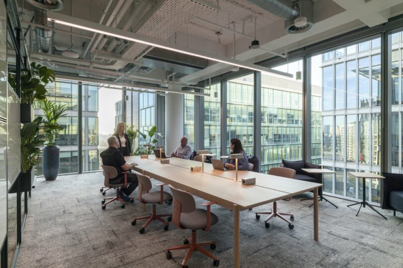 A modern office with large windows, a wooden conference table, and four people engaged in discussion. Virgin 02 green plants decorate the room, adding a fresh touch as laptop computers rest on the table.